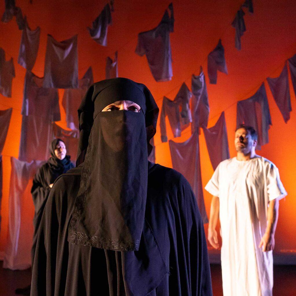 Two women in a black hijab and niqab, and a man in white traditional clothing in front of an orange backdrop and handing laundry,  in the stage play The Last 15 Seconds.