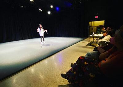Spectators watch a woman in white and black speaking at MT Space's TELL at The Registry Theatre in February of 2023.