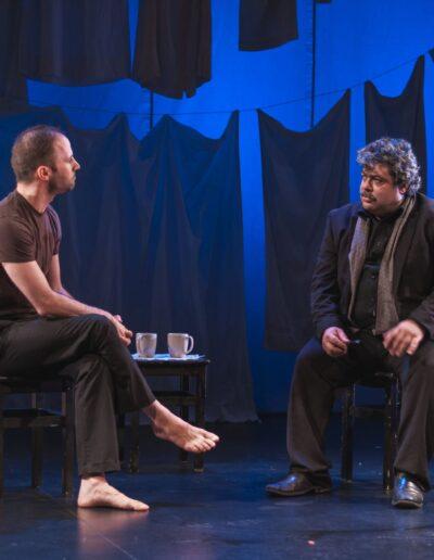 Trevor and Majdi in character for Last 15 Seconds. Both are staring at each other while sitting on stools, conducting an interview, against a blue background, with laundry draping down