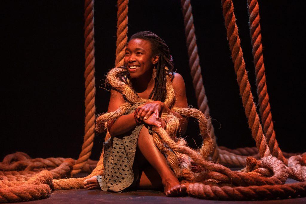 A woman, Shayna Jones, sits on the ground smiling with ropes hanging from the ceiling