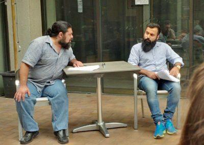 Majdi and Ahmad sit at a outdoor table with script in hand, both wearing grey shirts and blue jeans