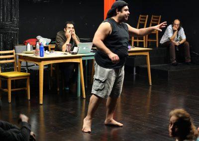 Majdi in shorts and tank top standing in middle of room directing actors, while Gary sits at table behind him, resting his chin on his hands