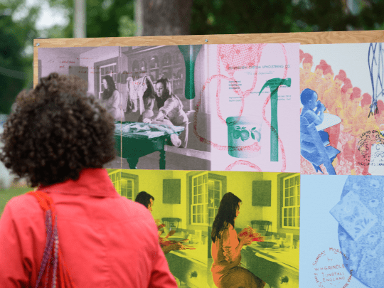 The Quiet Power of Personal Things A woman in a red blouse stands looking at an installation with a family portrait on it a map and various photos