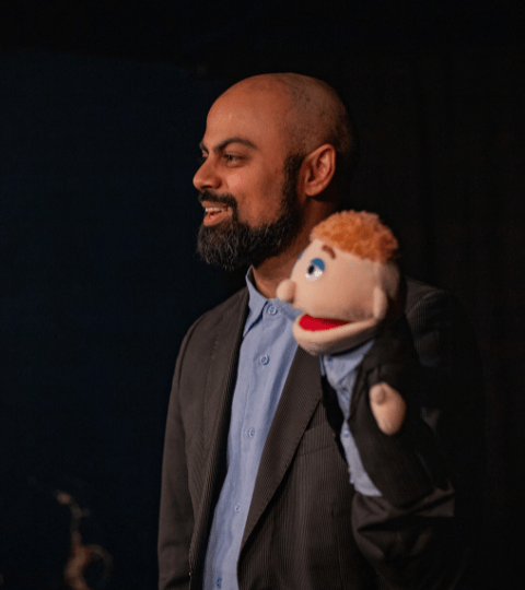 A man with a beard stands in a blue dress shirt and a sports coat. He carries a puppet with ginger wearing the same outfit as him