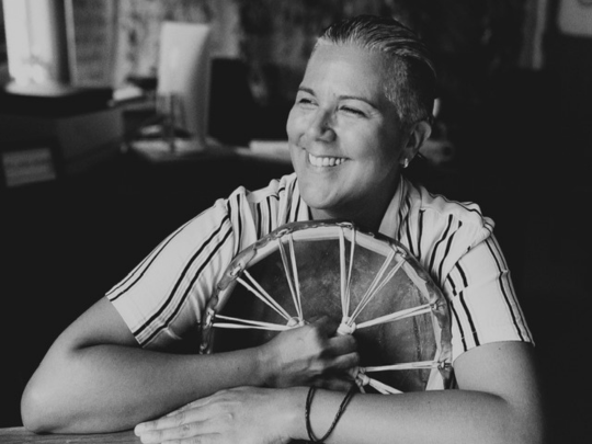Ni Nagamoon_ A Cultural Sharing Workshop - 540x405 A black and white photo of a woman holding a wooden wheel, smiling while looking into the distance