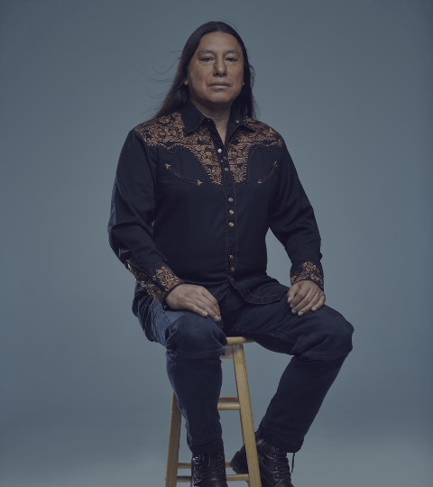 Indigenous man sitting on a wooden stool with black jeans on and a button up with golden accents