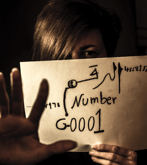 A woman holds a paper with "Number G0001" written on it in arabic. The paper covers her face and only her blonde hair peaks over the top. She holds one hand Infront of the paper signaling stop