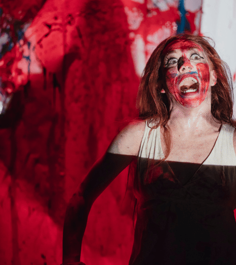 A woman screaming in the foreground with her face covered in red paint. She is in a black dress. The background is a white cloth covered in red paint