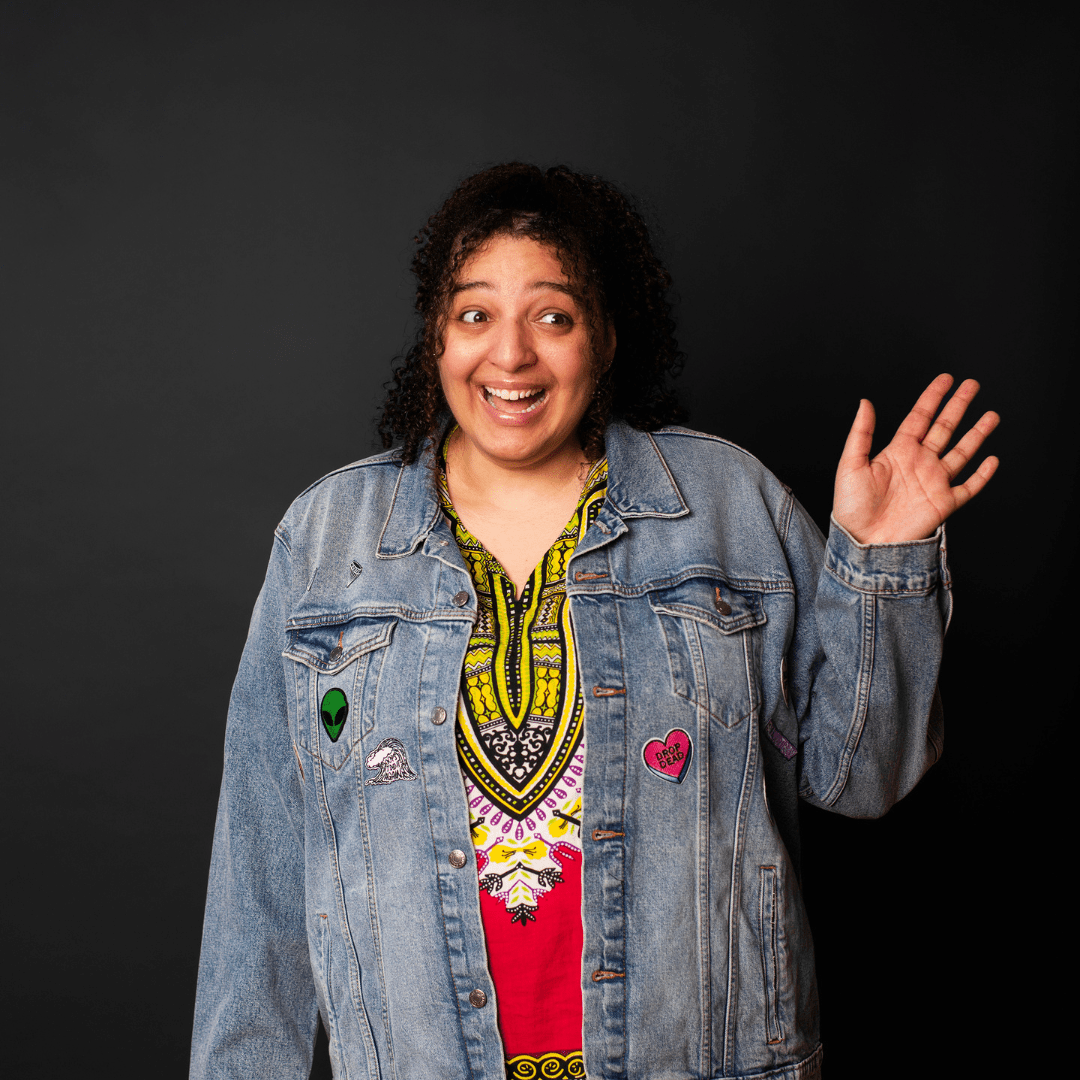 Sam, a biracial woman, wearing a dashiki with jean jacket over top. She has an awkward smile and waving to someone,